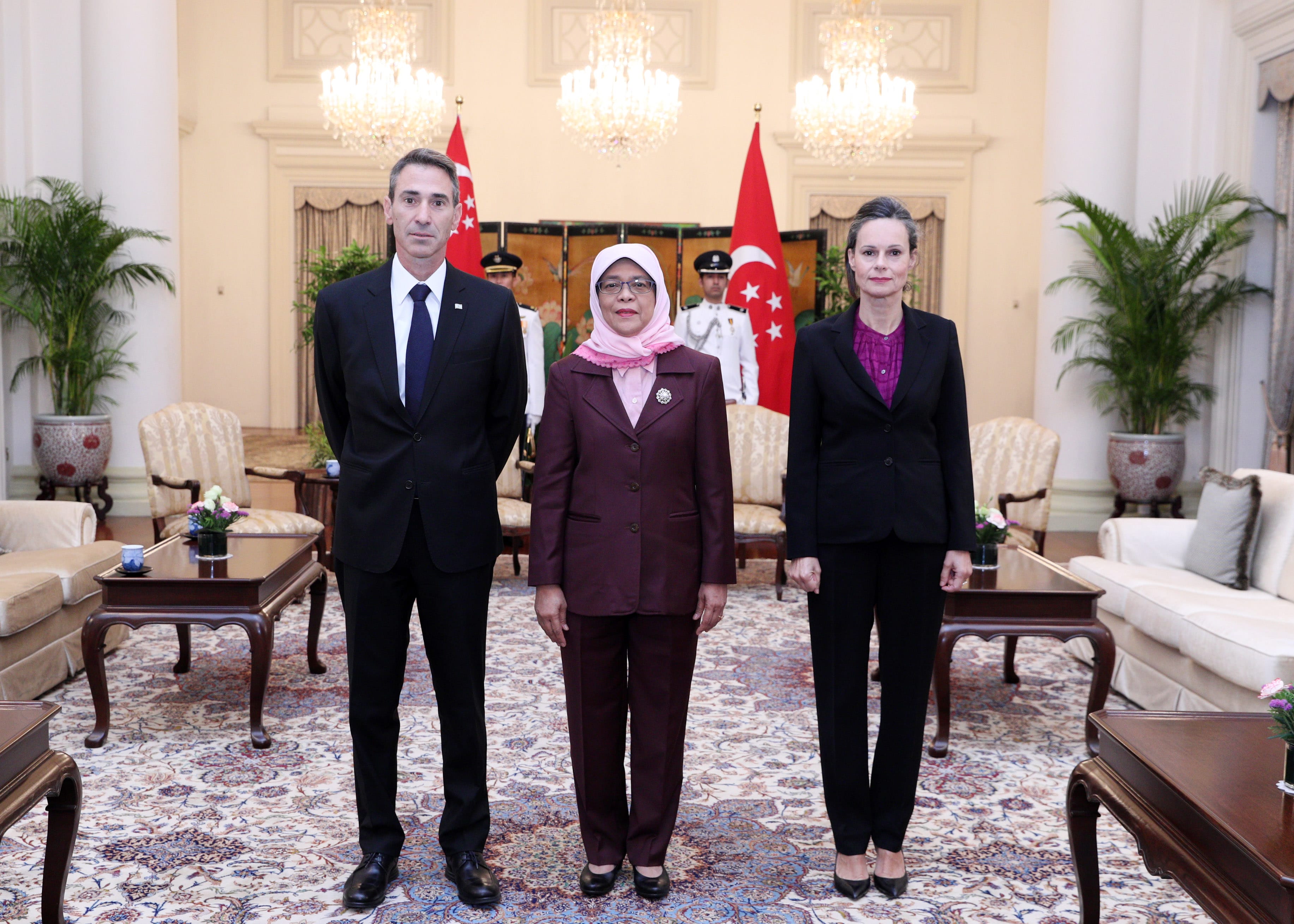 Three people in formal attire stand in a room with Singapore flags behind them.